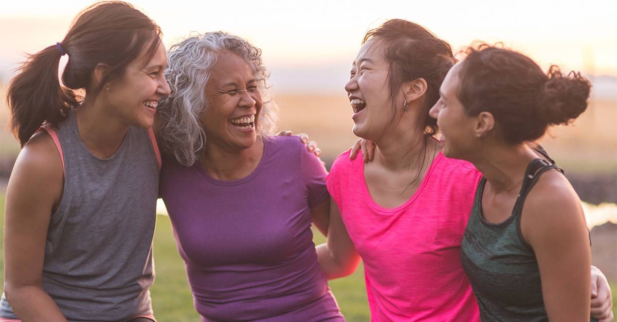 Four women with arms around each other, wearing casual workout clothes, standing outdoors during sunset.