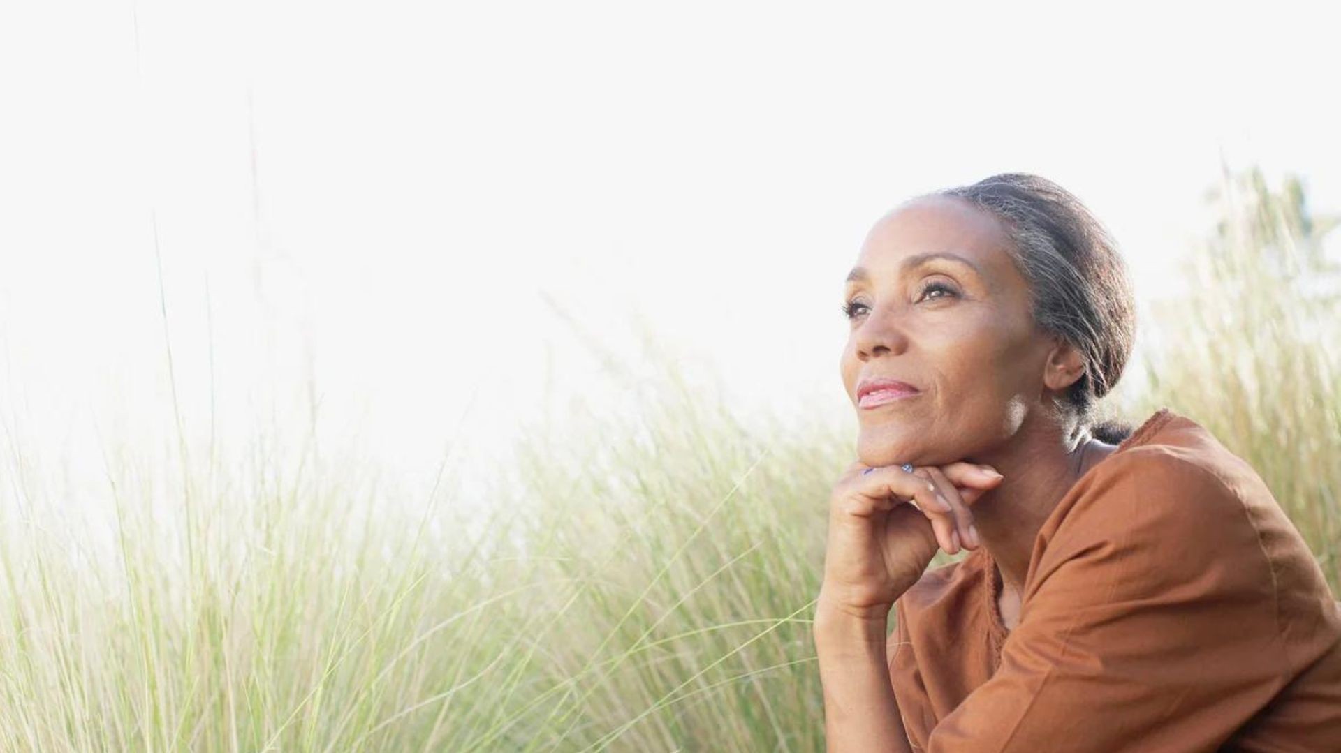 Person in a brown outfit sitting outdoors with tall grass in the background.