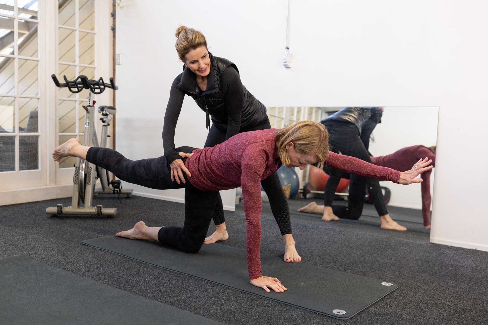 Instructor assisting woman in yoga pose on mat, with exercise equipment in the background.